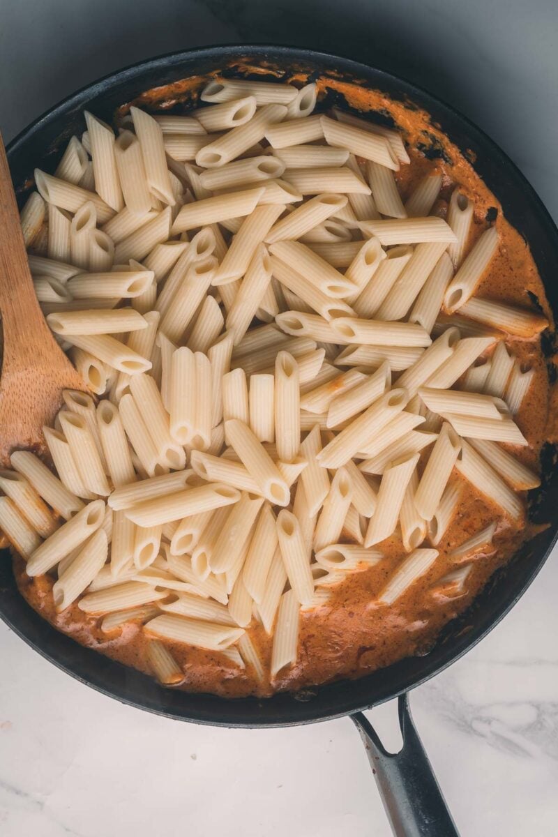 Cooked penne pasta sitting on top of creamy sauce in a black skillet with a wooden spoon on the left side.