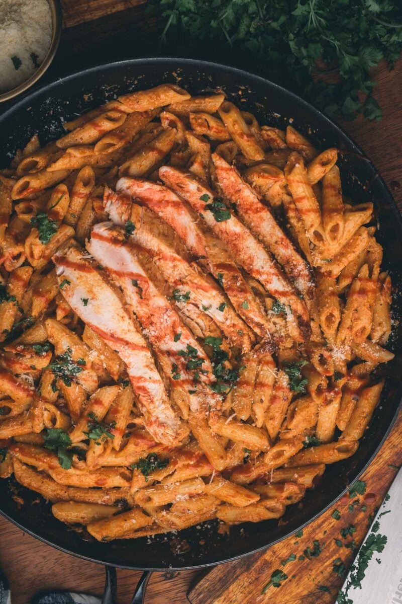 A skillet filled with penne pasta in a creamy orange sauce, topped with sliced grilled chicken breast and garnished with chopped parsley.