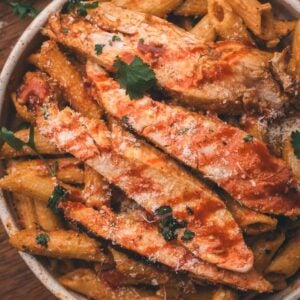 Bowl of penne pasta topped with sliced chipotle chicken, tomato sauce, grated cheese, and parsley on a wooden surface.