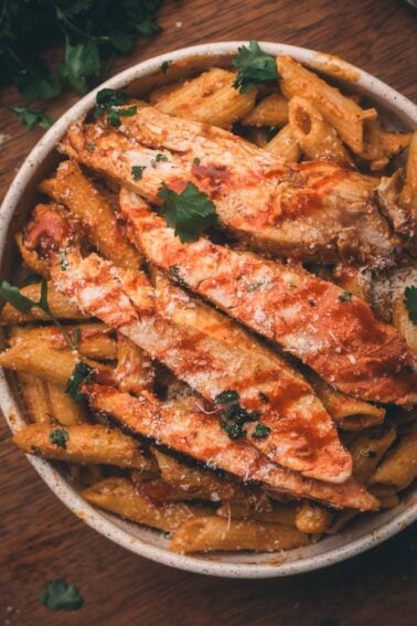 Bowl of penne pasta topped with sliced chipotle chicken, tomato sauce, grated cheese, and parsley on a wooden surface.