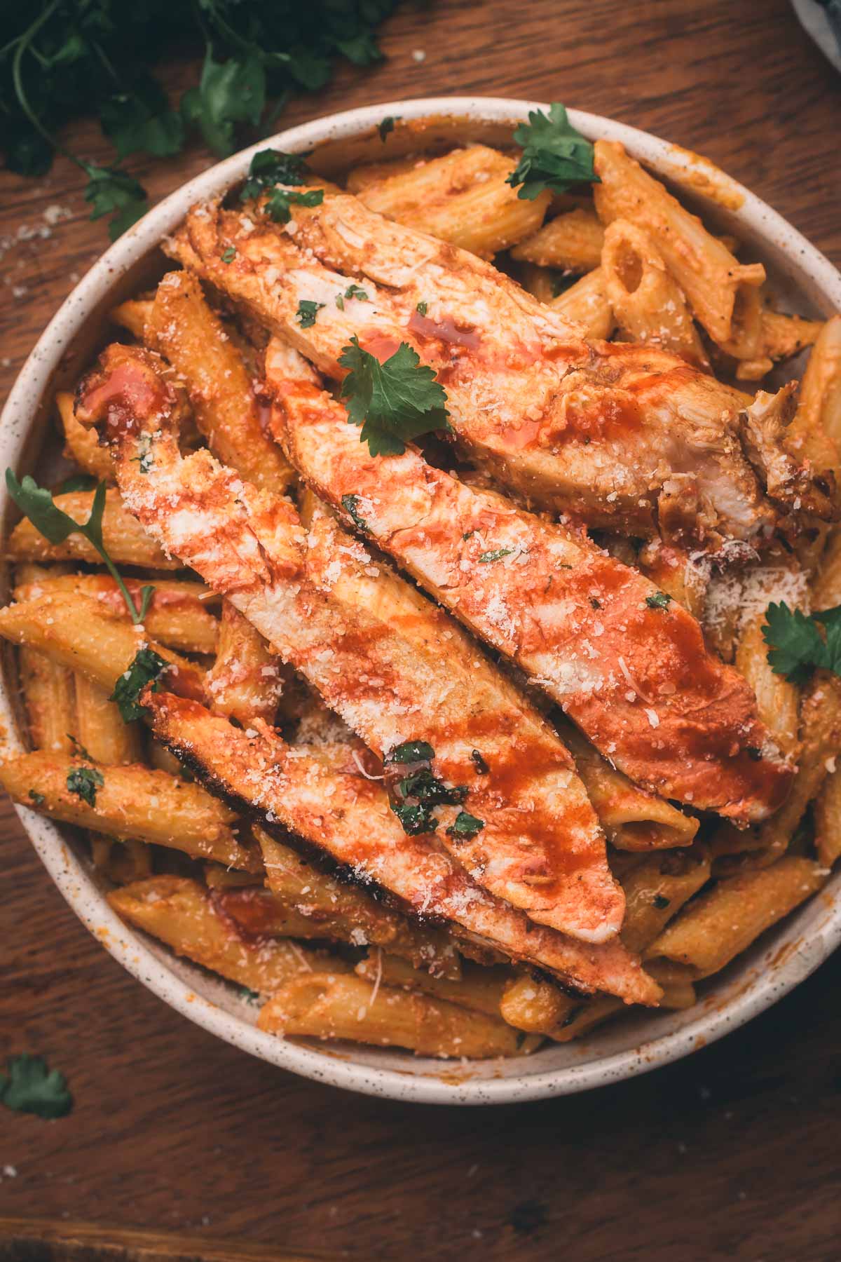 Bowl of penne pasta topped with sliced grilled chicken, garnished with grated cheese and parsley on a wooden surface.