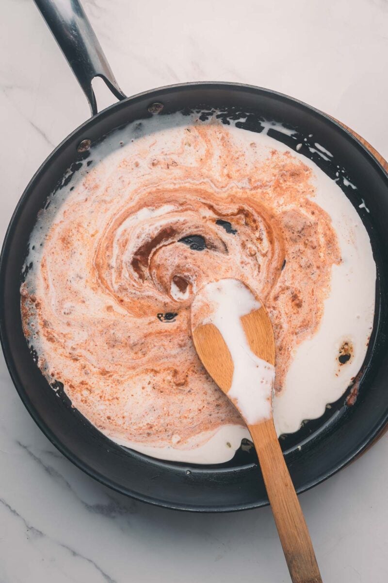 A wooden spoon stirs cream and spices in a black skillet on a marble surface.