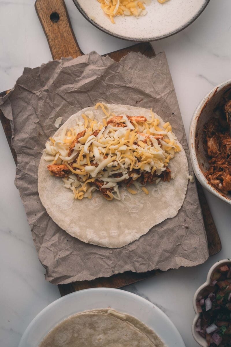 A tortilla on parchment paper topped with shredded chicken and cheese, with bowls of ingredients visible nearby on a white surface.