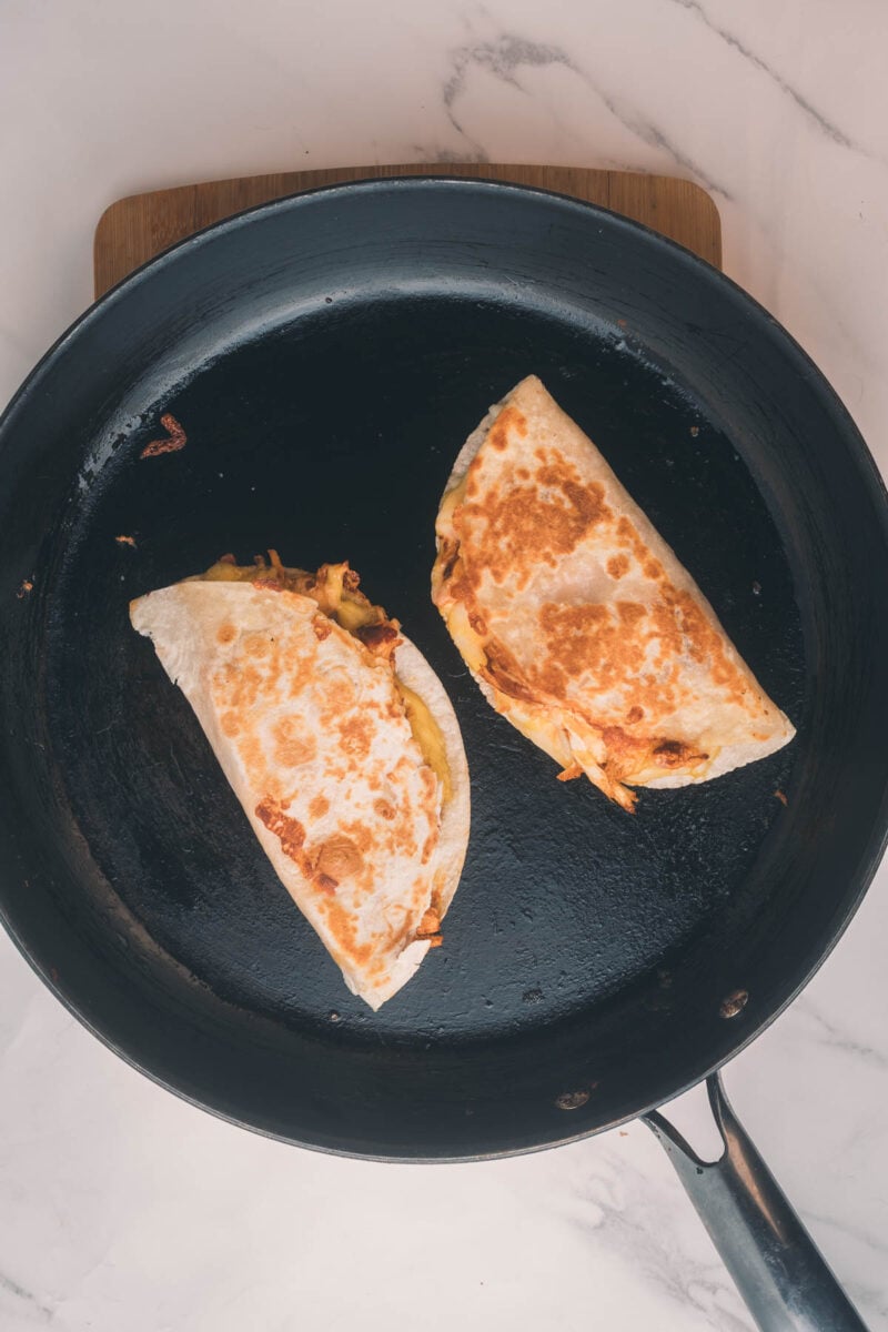 Two folded tortillas with melted cheese and fillings are cooking in a black skillet on a marble countertop.