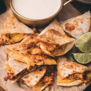 Chicken quesadilla wedges arranged on parchment paper with a cup of white dipping sauce and lime slices on the side.