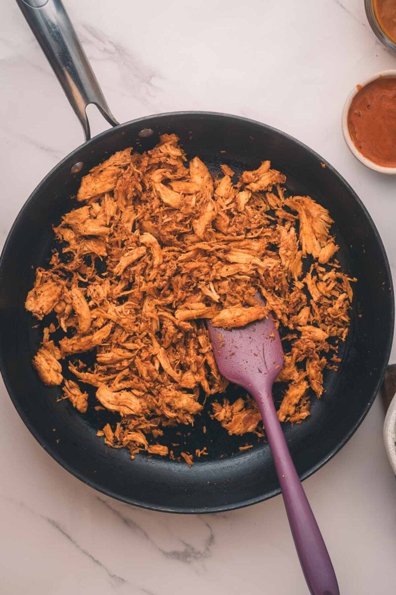 Shredded chipotle chicken cooking in a black skillet with a purple spatula, on a white countertop with sauce bowls nearby.