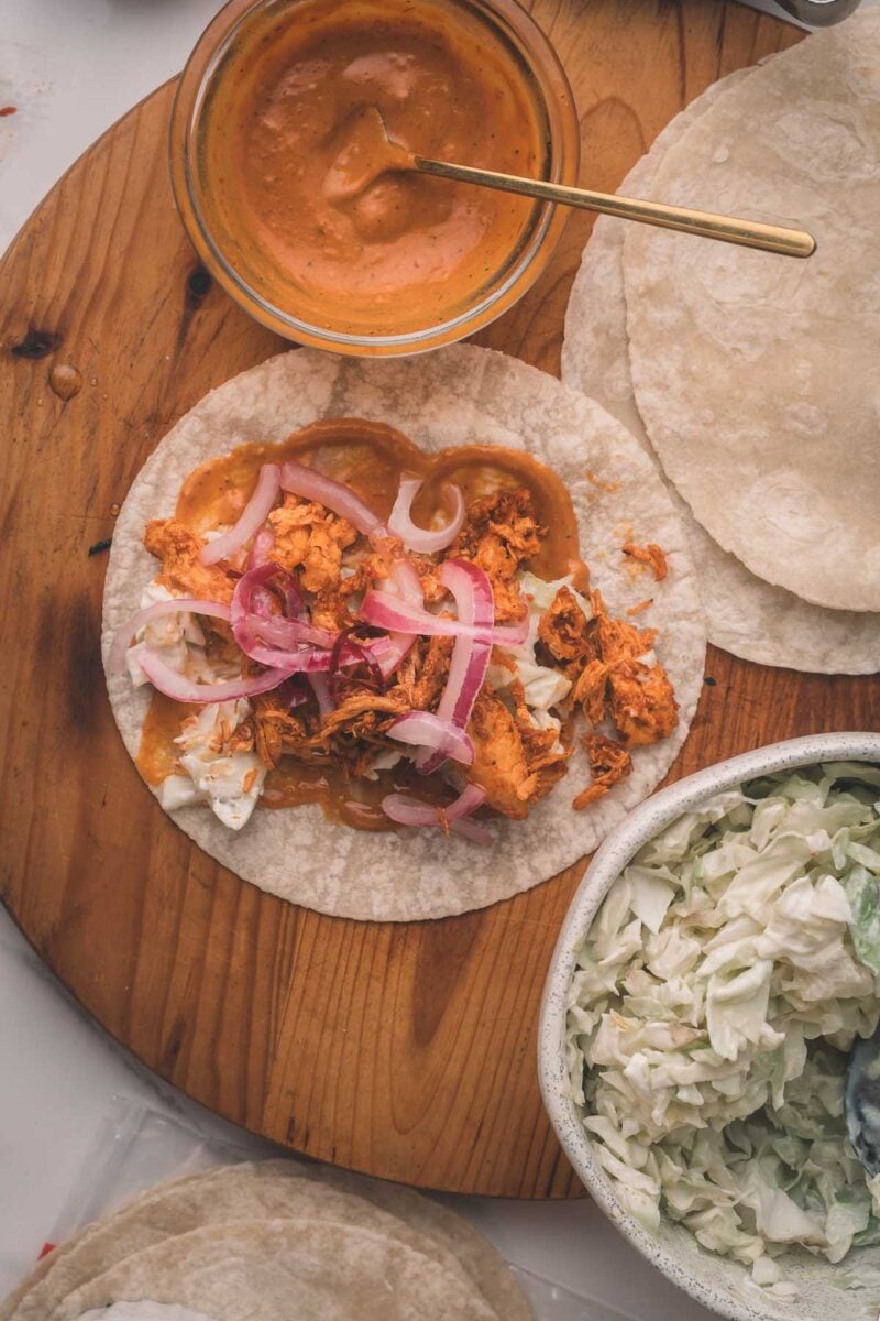 Open tortilla topped with shredded chicken, pickled red onions, and slaw on a wooden board, with a bowl of creamy sauce and extra slaw nearby.