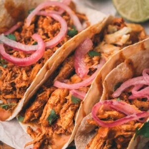 Three shredded chipotle chicken tacos topped with pickled red onions and cilantro, served on soft tortillas with lime wedges in the background.