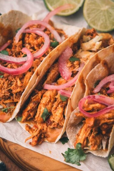 Three shredded chiptole chicken tacos topped with pickled red onions and cilantro, served on soft tortillas with lime wedges in the background.