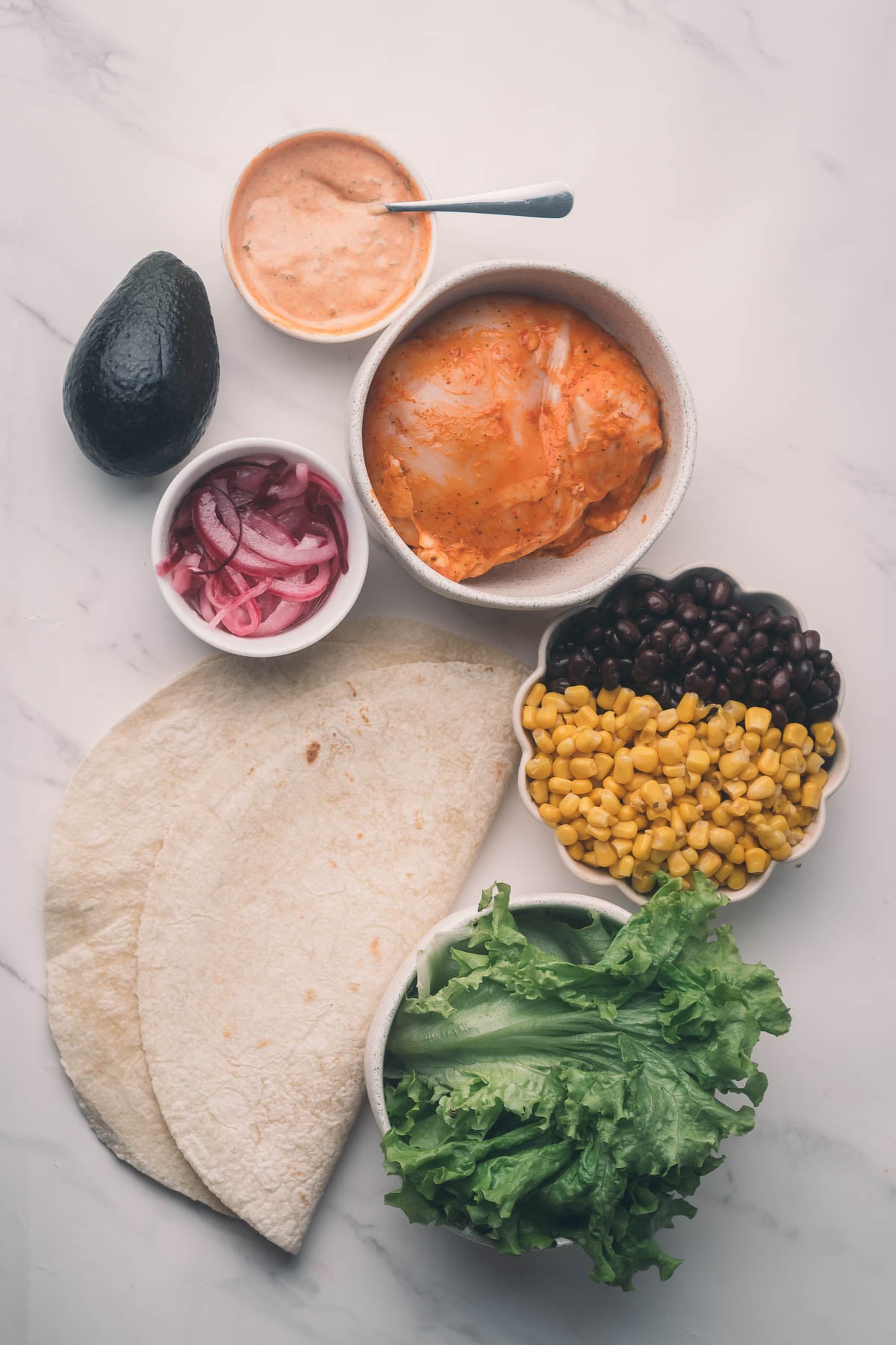 Ingredients for chipotle chicken wraps arranged on a counter: tortillas, lettuce, corn, black beans, avocado, pickled onions, marinated chicken, and a bowl of sauce with a spoon.