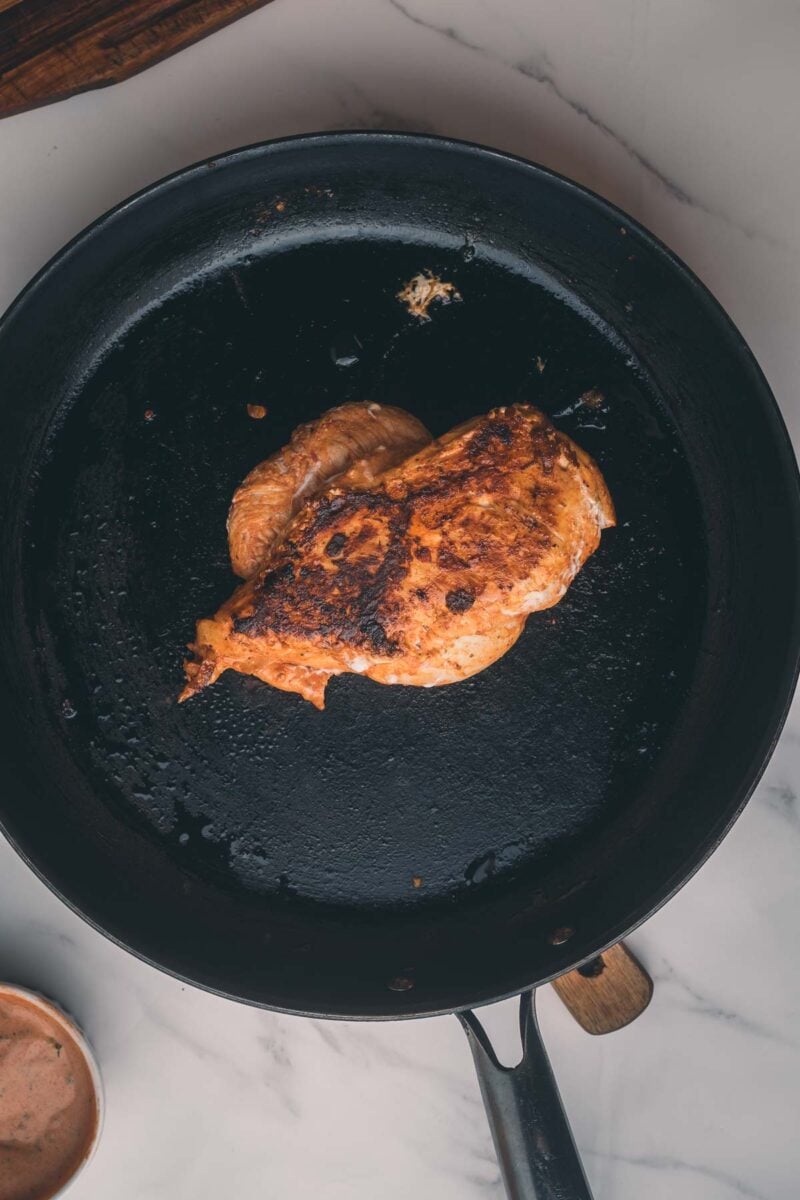 A cooked, seasoned chicken breast sits in a black skillet on a white marble countertop.