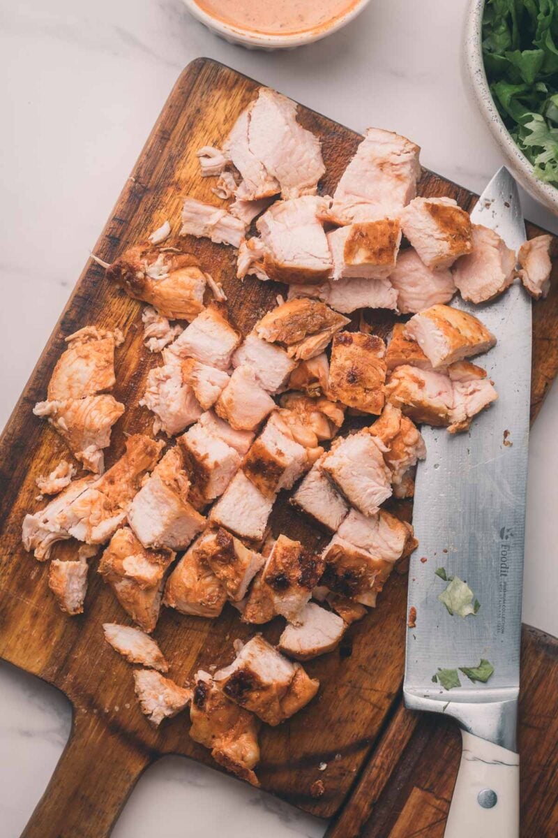 Cooked chicken breast cut into bite-sized pieces on a wooden cutting board, with a large knife and some chopped cilantro nearby.