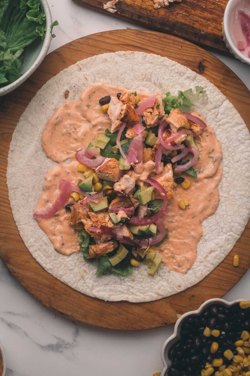 A tortilla on a wooden board topped with lettuce, chopped chipotle chicken, corn, black beans, avocado, pickled onions, and creamy sauce, ready to be wrapped.