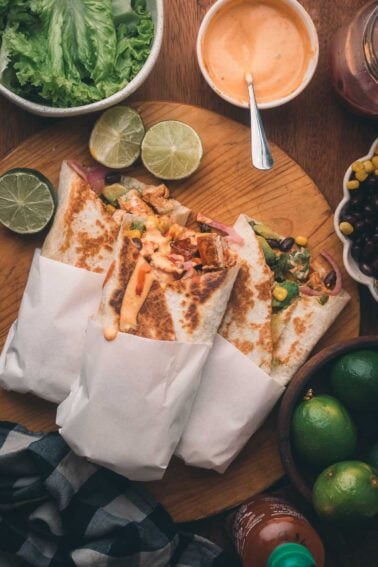 Three crispy chipotle chicken wraps in paper on a wooden board, topped with sauce, surrounded by limes, lettuce, black beans, and a bowl of orange dipping sauce.