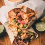 Two chicken wraps with corn, avocado, onions, and sauce are cut in half on a wooden board, with lime wedges and a bowl of orange sauce nearby.