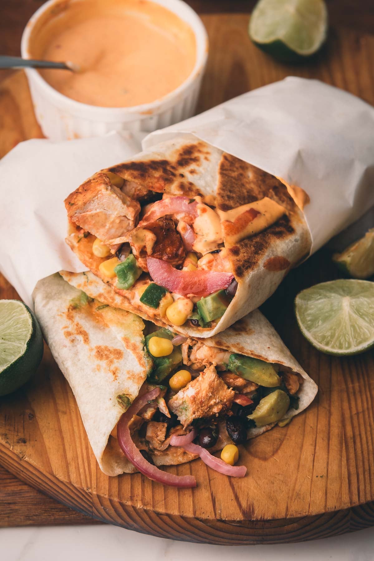 Two chicken wraps with corn, avocado, onions, and sauce are cut in half on a wooden board, with lime wedges and a bowl of orange sauce nearby.
