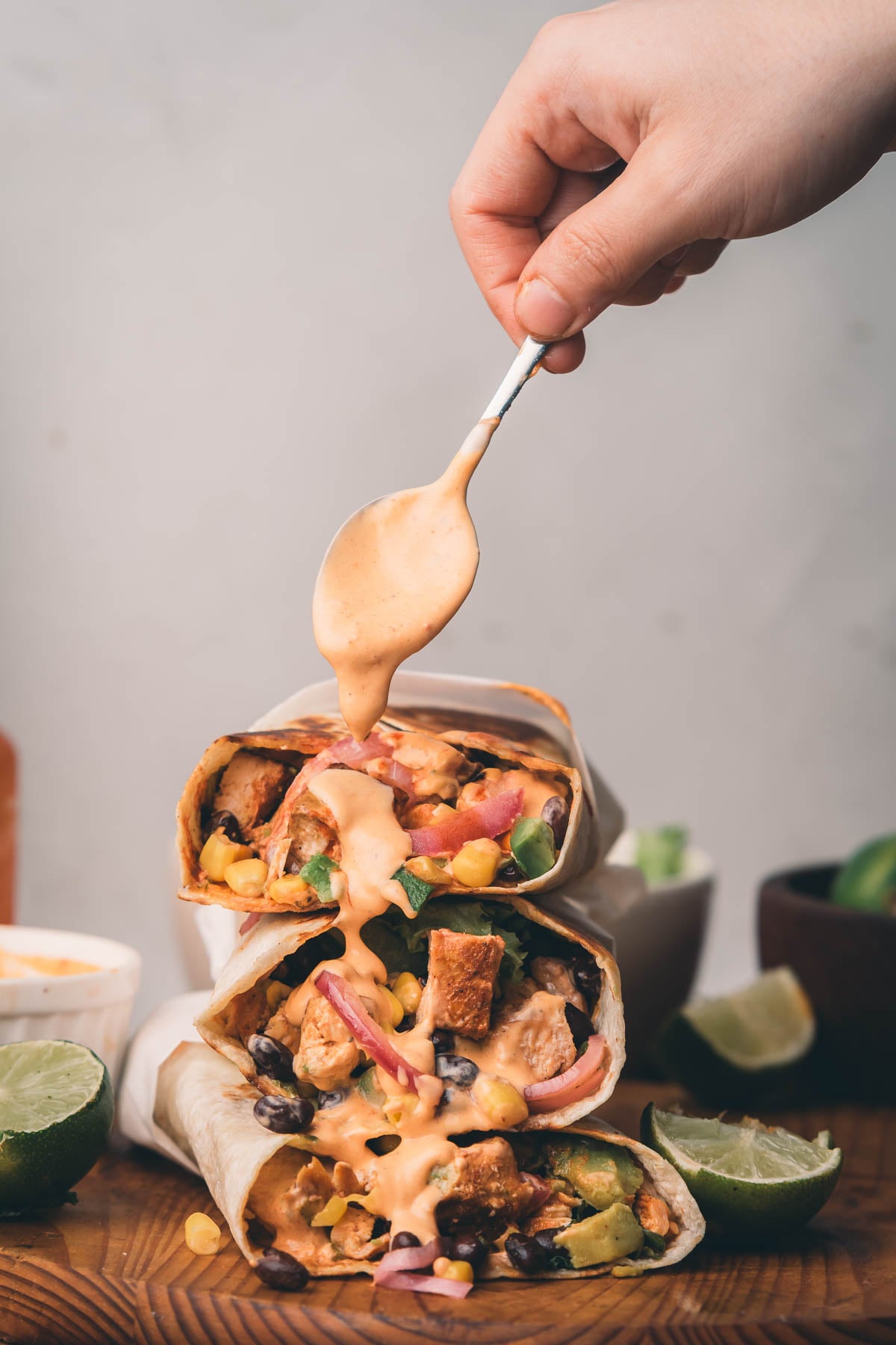 A hand drizzles sauce over three stacked wraps filled with beans, corn, chipotle chicken, and vegetables. Lime halves and a bowl of sauce are on the wooden surface.