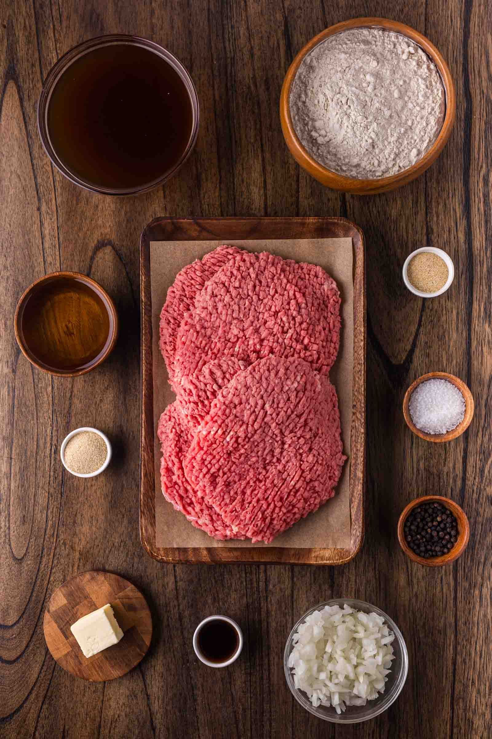Raw tenderized cube steaks on a tray surrounded by bowls of flour, chopped onions, butter, broth, spices, Worcestershire sauce, and seasonings, arranged on a wooden surface.