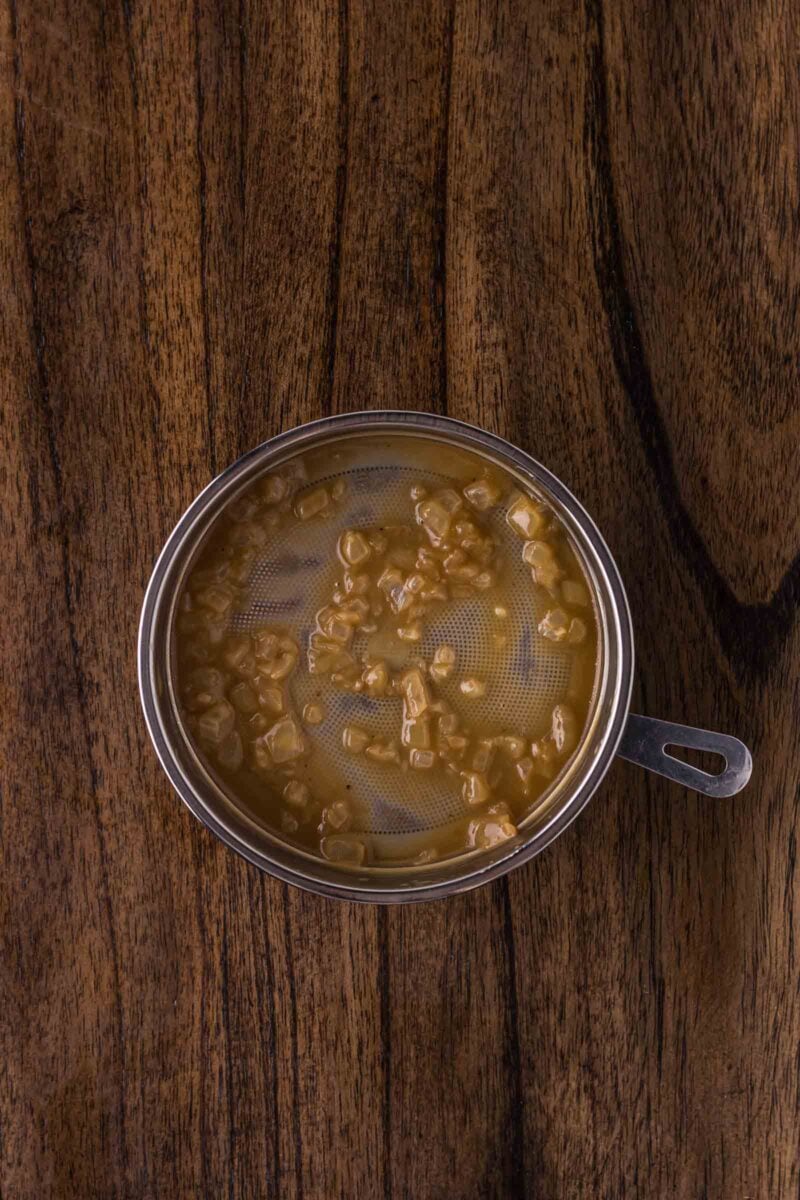 A metal strainer with diced onions in liquid is placed on a wooden surface.