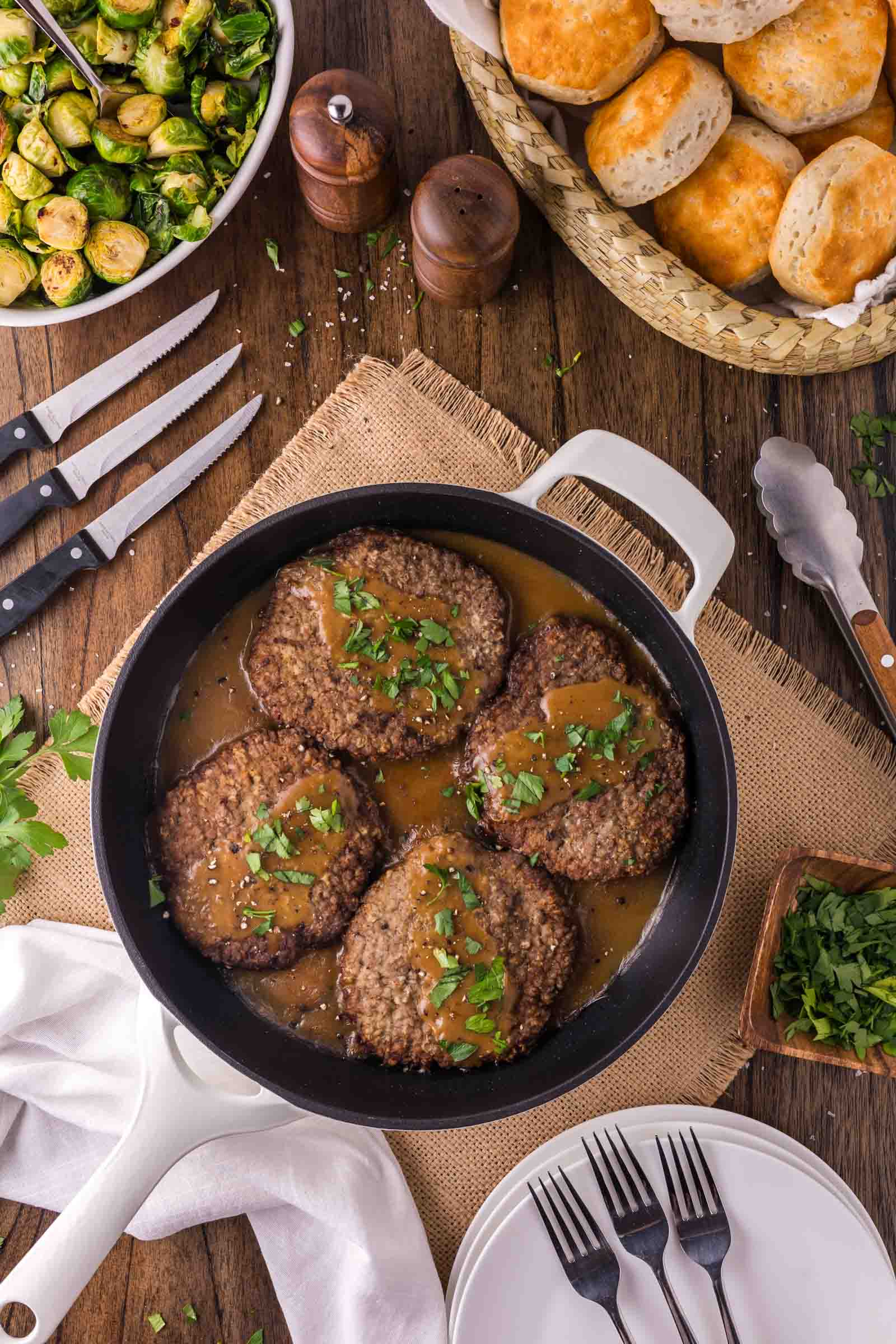 A skillet with country fried steaks in gravy, garnished with herbs, surrounded by Brussels sprouts, biscuits, and dining utensils on a wooden table.