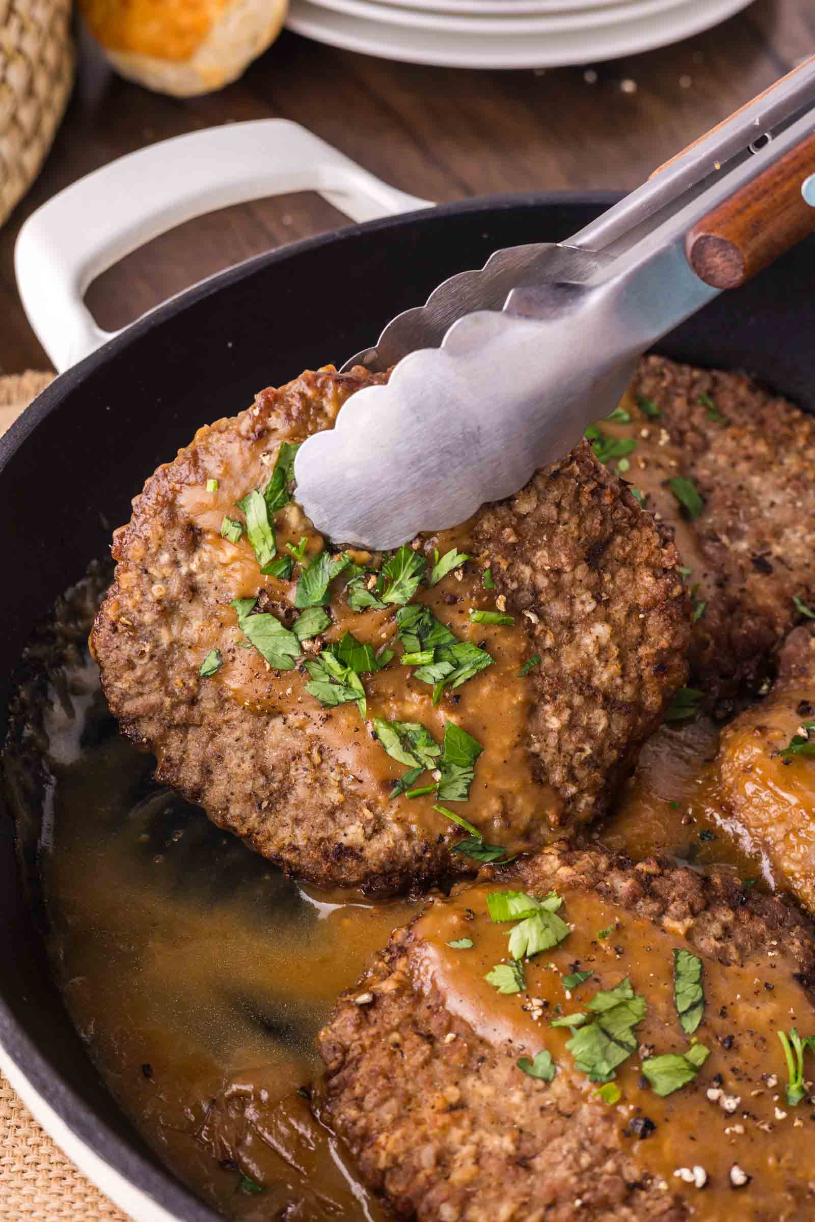 A pair of tongs lifts a country fried steak with brown gravy and chopped herbs from a skillet.
