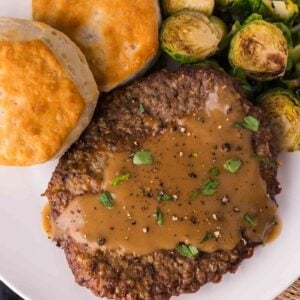 A plate with a country fried cube steak topped with brown gravy, roasted Brussels sprouts, and two biscuits.