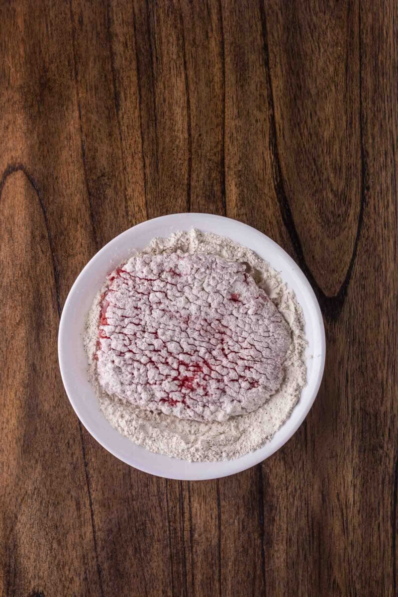 Raw cube steak coated in flour sits on a white plate, placed on a wooden surface.