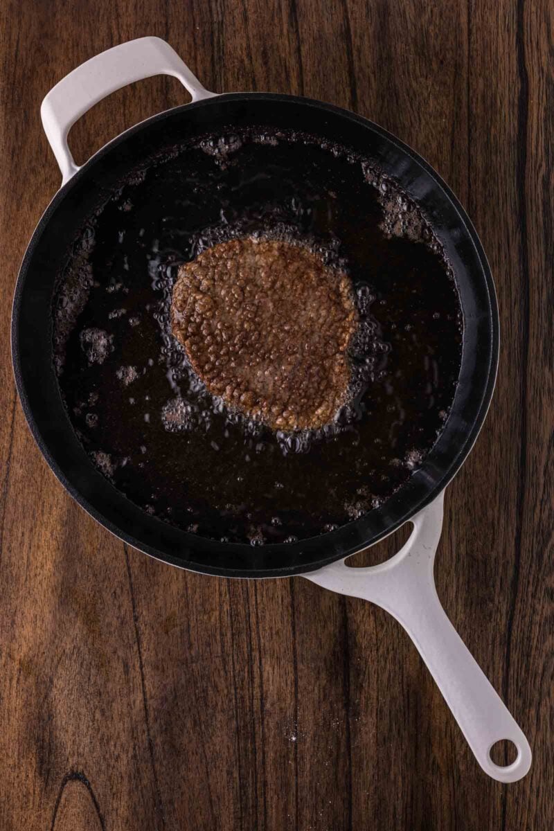 A round cube steak is deep frying in a black pan with a white handle, placed on a wooden surface.