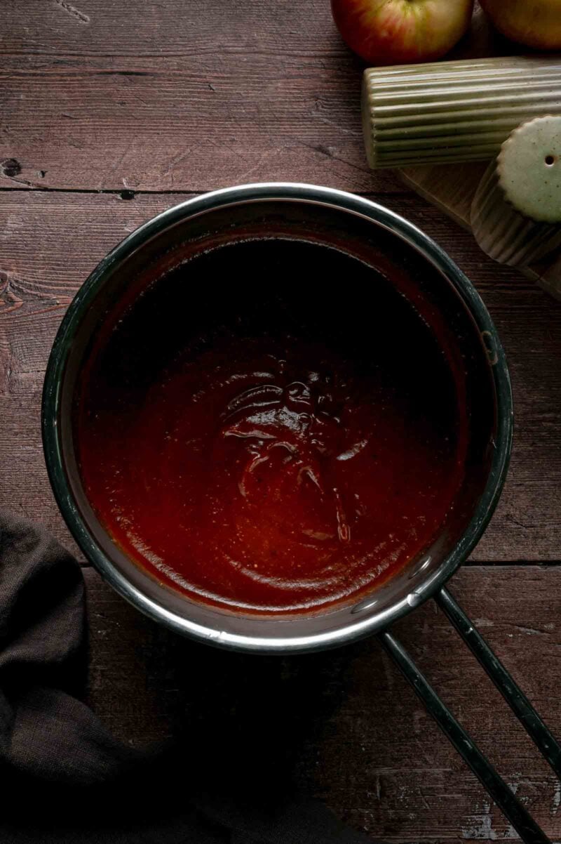 Apple butter BBQ sauce simmered smooth and glossy in the saucepan, thick enough to coat a wing, with apples in the background.