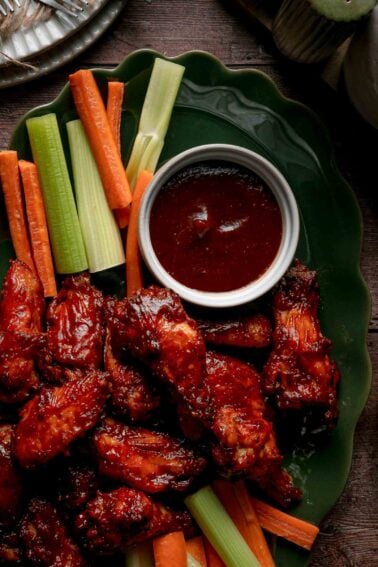 Close up of apple butter BBQ wings on a green platter beside celery, carrots and a ramekin of reserved apple butter BBQ sauce.