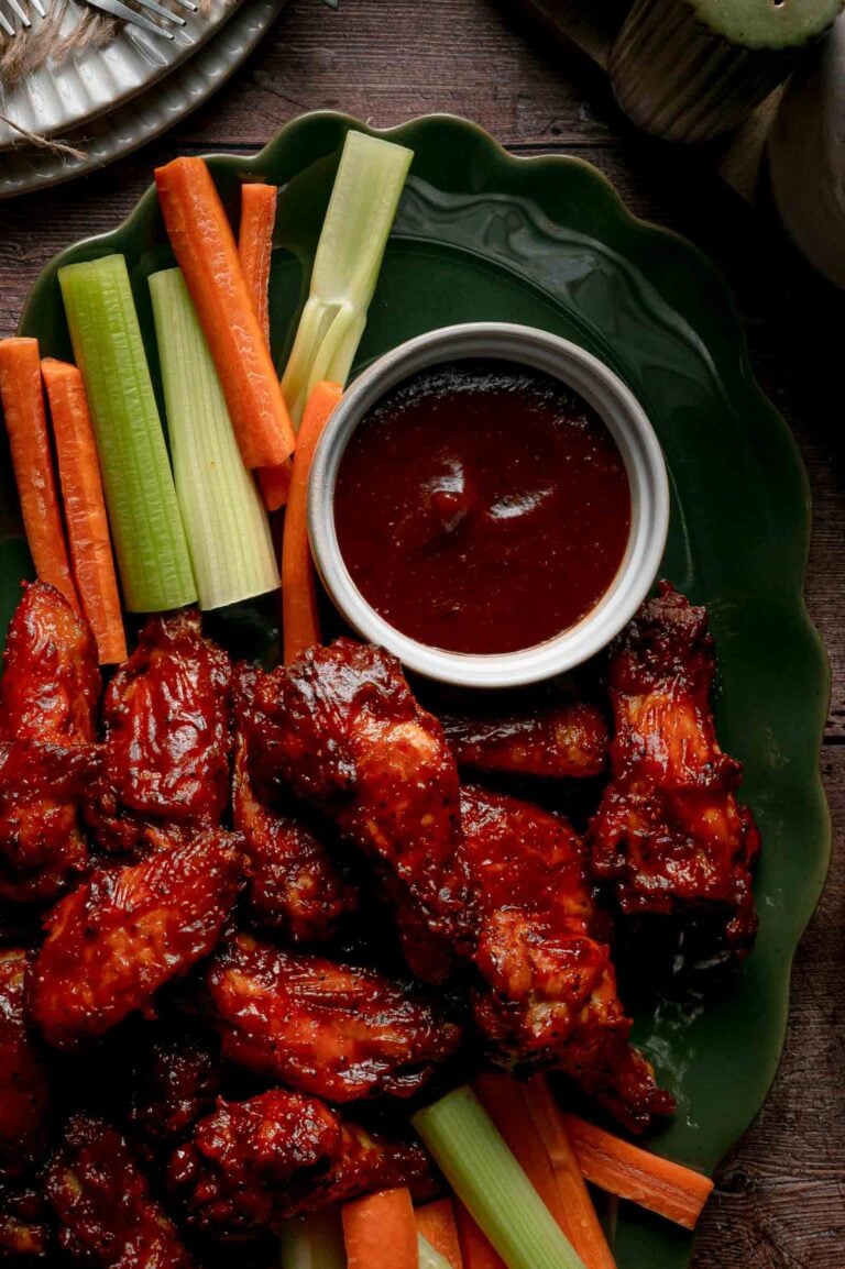 Close up of apple butter BBQ wings on a green platter beside celery, carrots and a ramekin of reserved apple butter BBQ sauce.
