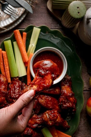 An apple butter BBQ wing lifted dripping from the ramekin of sauce, the rest of the loaded platter below.