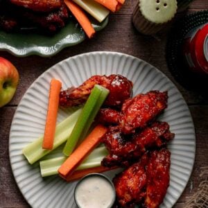 A plate of apple butter BBQ wings with carrot and celery sticks and a cup of creamy white dipping sauce, apples alongside.