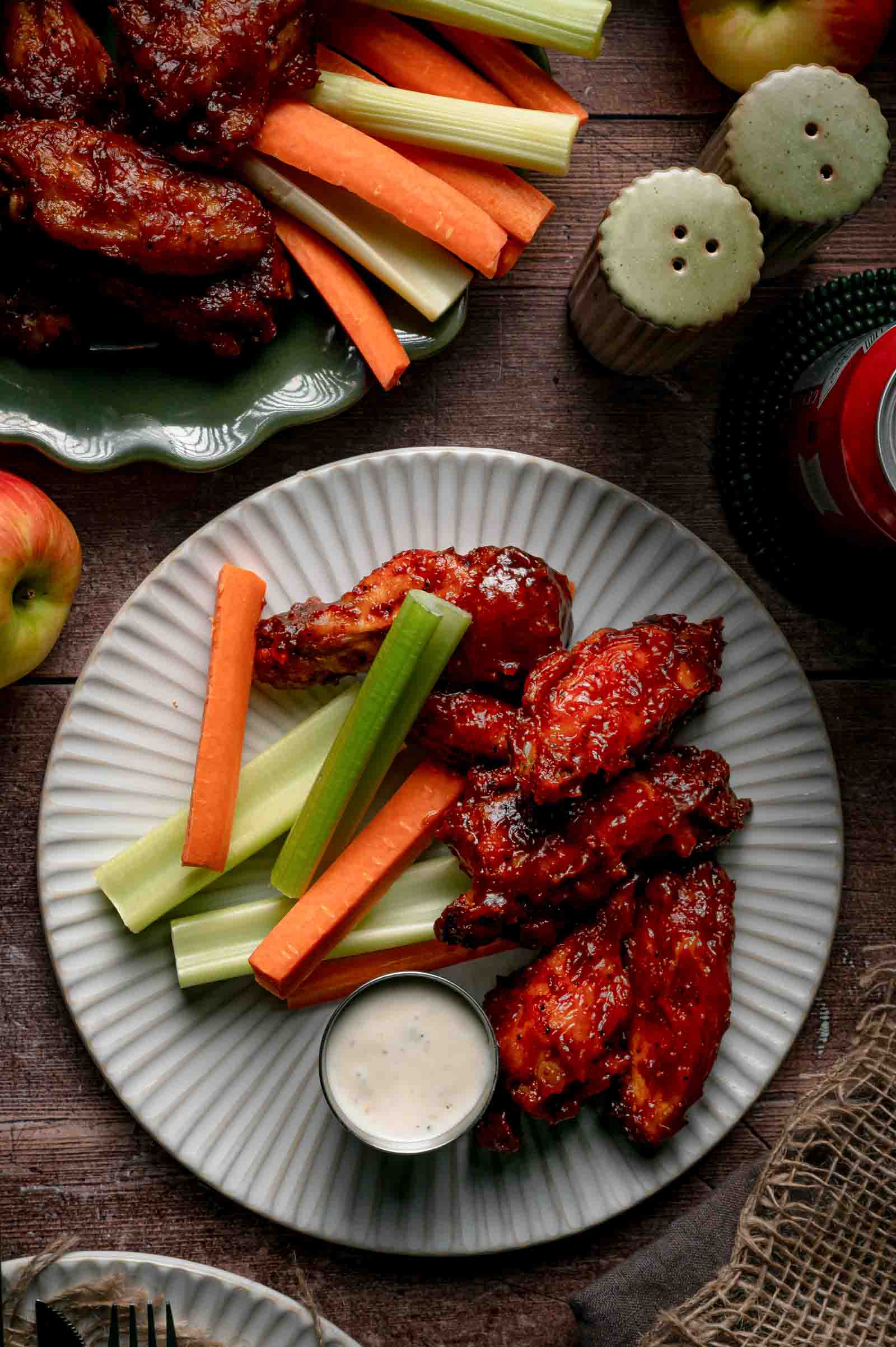 A plate of apple butter BBQ wings with carrot and celery sticks and a cup of creamy white dipping sauce, apples alongside.