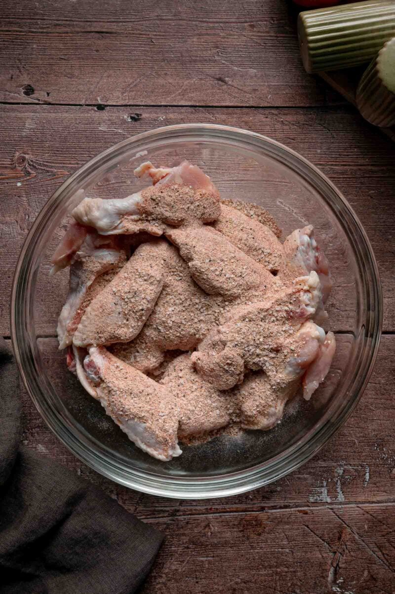 Raw chicken wings in a glass bowl with the baking powder seasoning blend dumped over the top, before tossing.