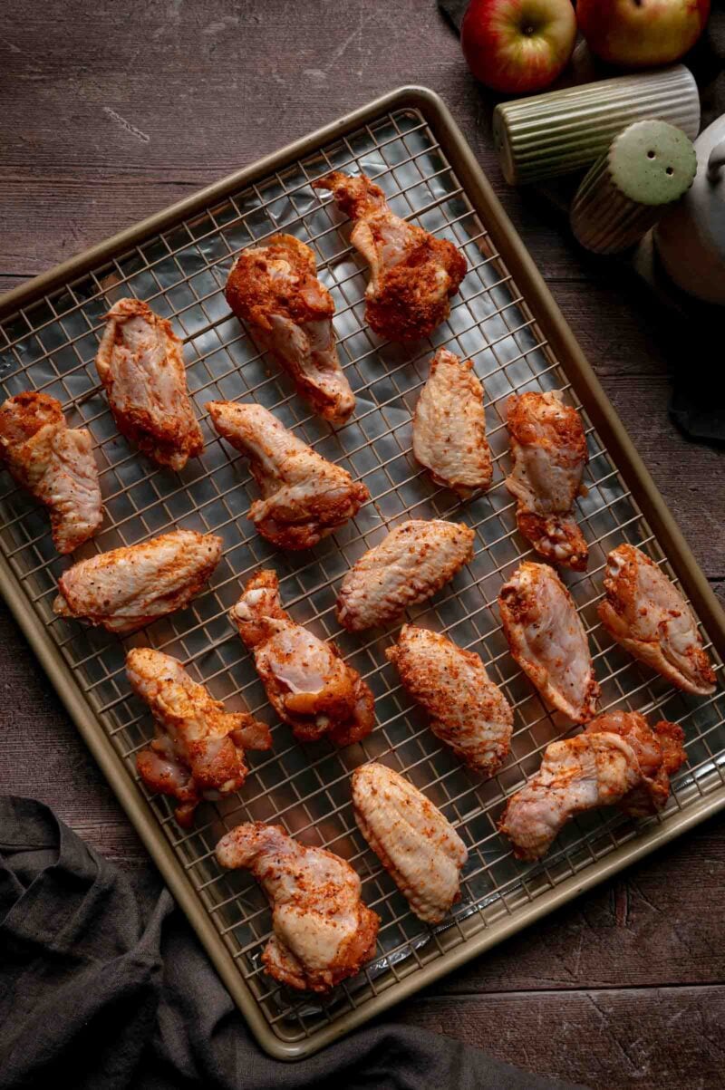 Seasoned raw chicken wings spaced out on a wire rack set over a foil-lined sheet pan, ready for the uncovered chill in the fridge.