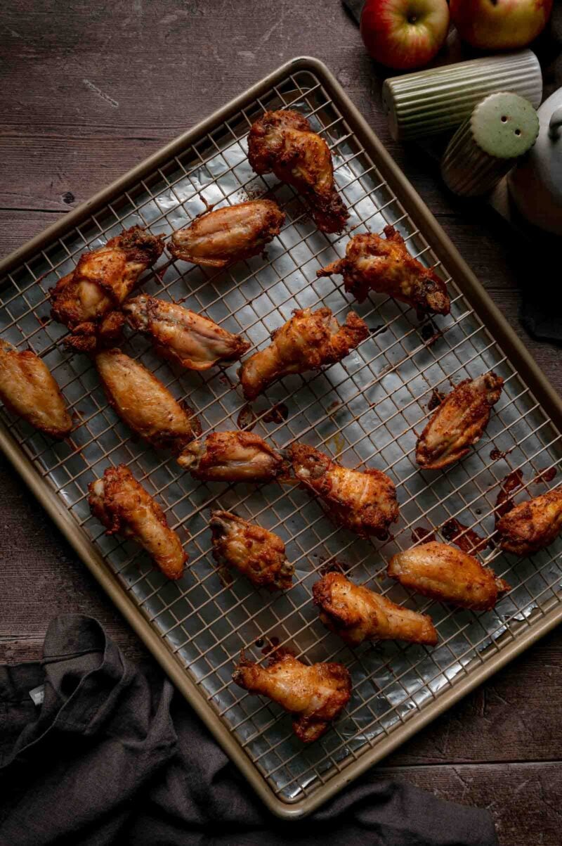 Chicken wings baked until the skin is deep golden and crisp, still unsauced on the wire rack, with apples in the background.