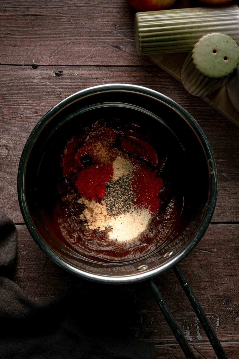 Apple butter, ketchup and brown sugar in a saucepan with paprika, black pepper, onion powder and garlic powder piled on top, before stirring.