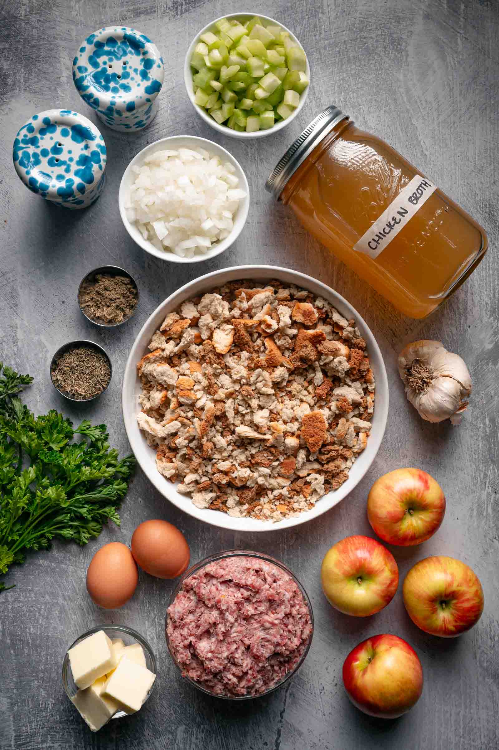 Ingredients for apple sausage stuffing: stuffing cubes, raw sage pork sausage, Honeycrisp apples, celery, onion, garlic, eggs, butter, parsley and a jar of chicken broth.
