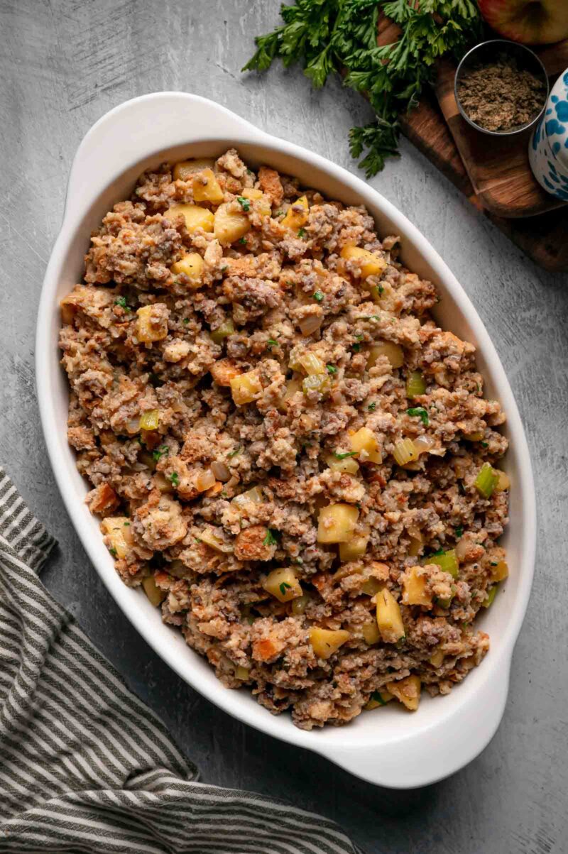The unbaked apple sausage stuffing spooned loosely into a white oval baking dish before it goes in the oven.