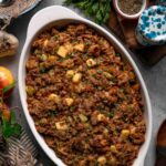 Baked apple sausage stuffing in a white oval dish, browned on top and scattered with parsley, surrounded by apples and herbs.