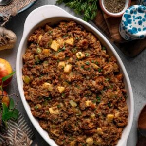 Baked apple sausage stuffing in a white oval dish, browned on top and scattered with parsley, surrounded by apples and herbs.
