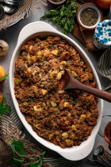 A wooden spoon lifting a scoop from a dish of baked apple sausage stuffing, showing the soft center under the crisp top.