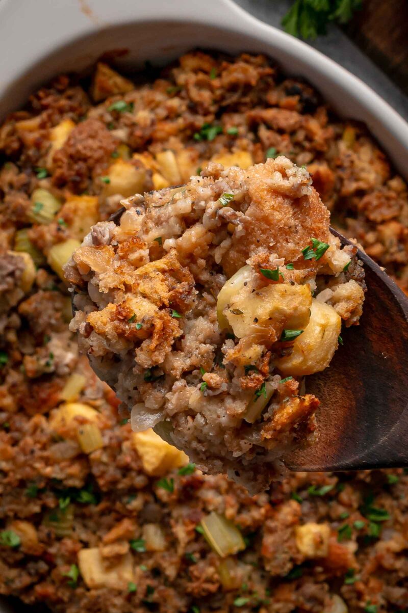 A wooden spoon holding a serving of apple sausage stuffing above the baking dish, crisp edges and apple chunks in view.