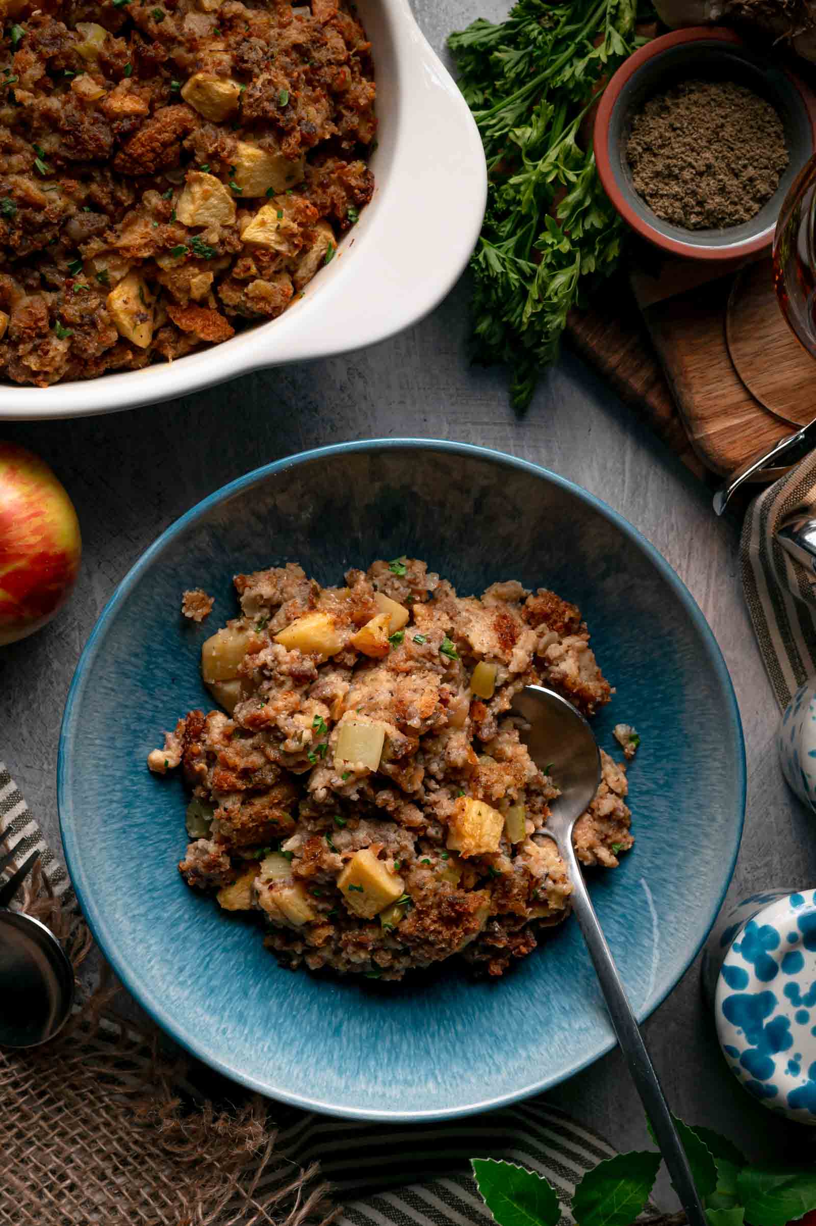 A blue bowl of apple sausage stuffing served beside the casserole dish, with a spoon, apples and fresh herbs on the table.