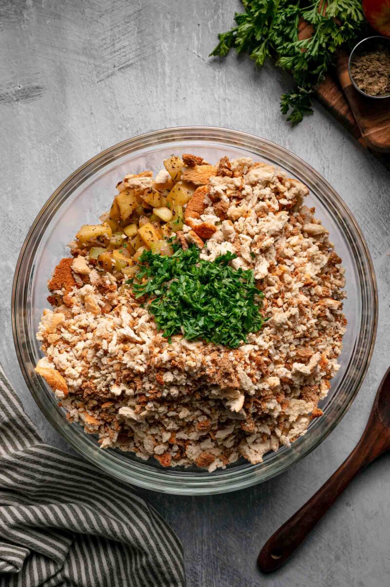 Stuffing cubes in a glass bowl with the cooked apple and vegetable mixture and a pile of chopped fresh parsley.