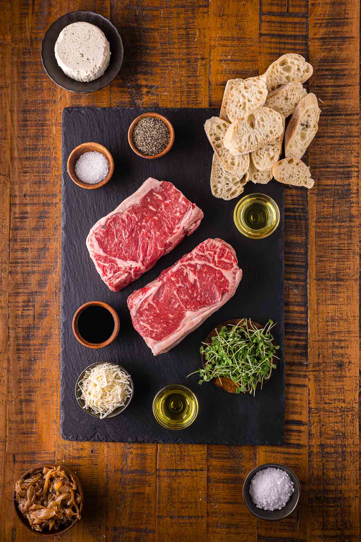 Ingredients for steak crostini: raw NY strip steaks, a round of garlic herb Boursin, baguette slices, caramelized onions, balsamic and Parmesan.