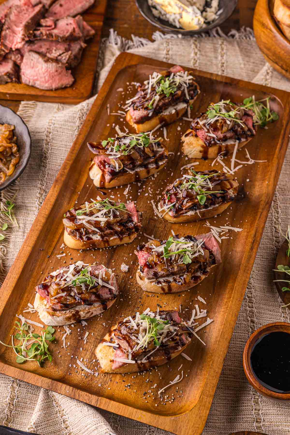 A wood tray of eight steak crostini with Boursin, caramelized onions, shaved Parmesan and microgreens, balsamic glaze drizzled over.
