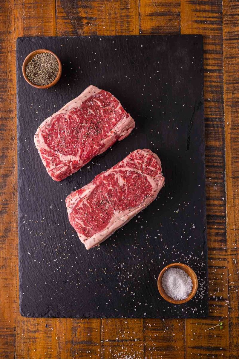 Two raw NY strip steaks seasoned with kosher salt and cracked black pepper on a slate board, resting before the sear.