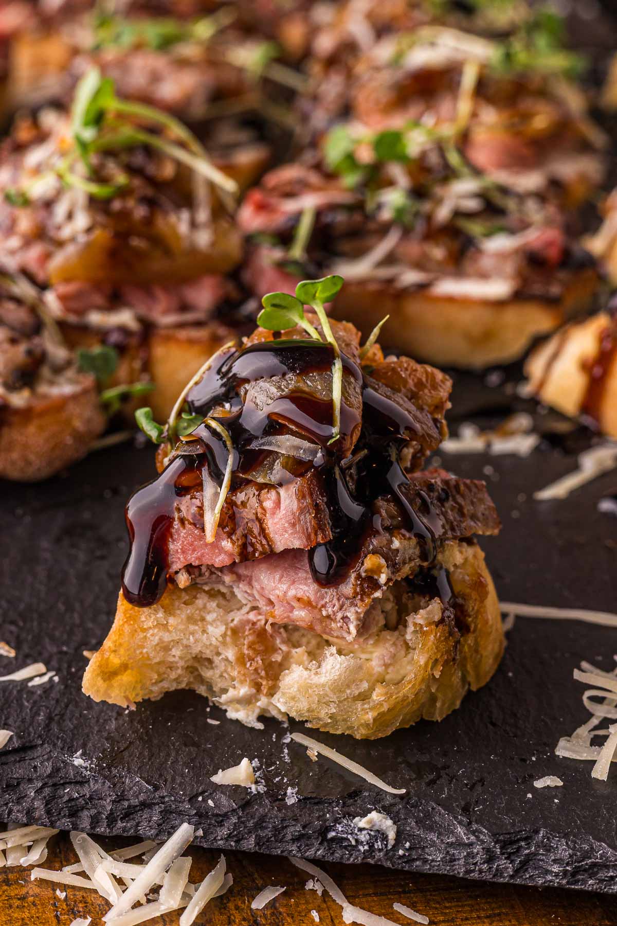 A steak crostini with a bite taken out of it, standing on slate with balsamic glaze running over the steak and onions.